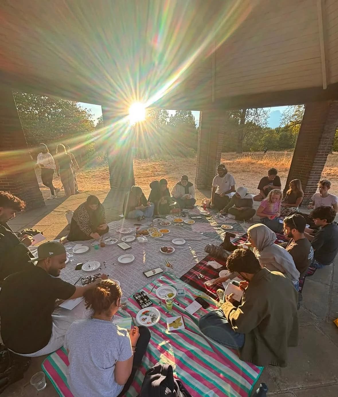 A group sitting on picnic blankets under a park shelter, painting at golden hour